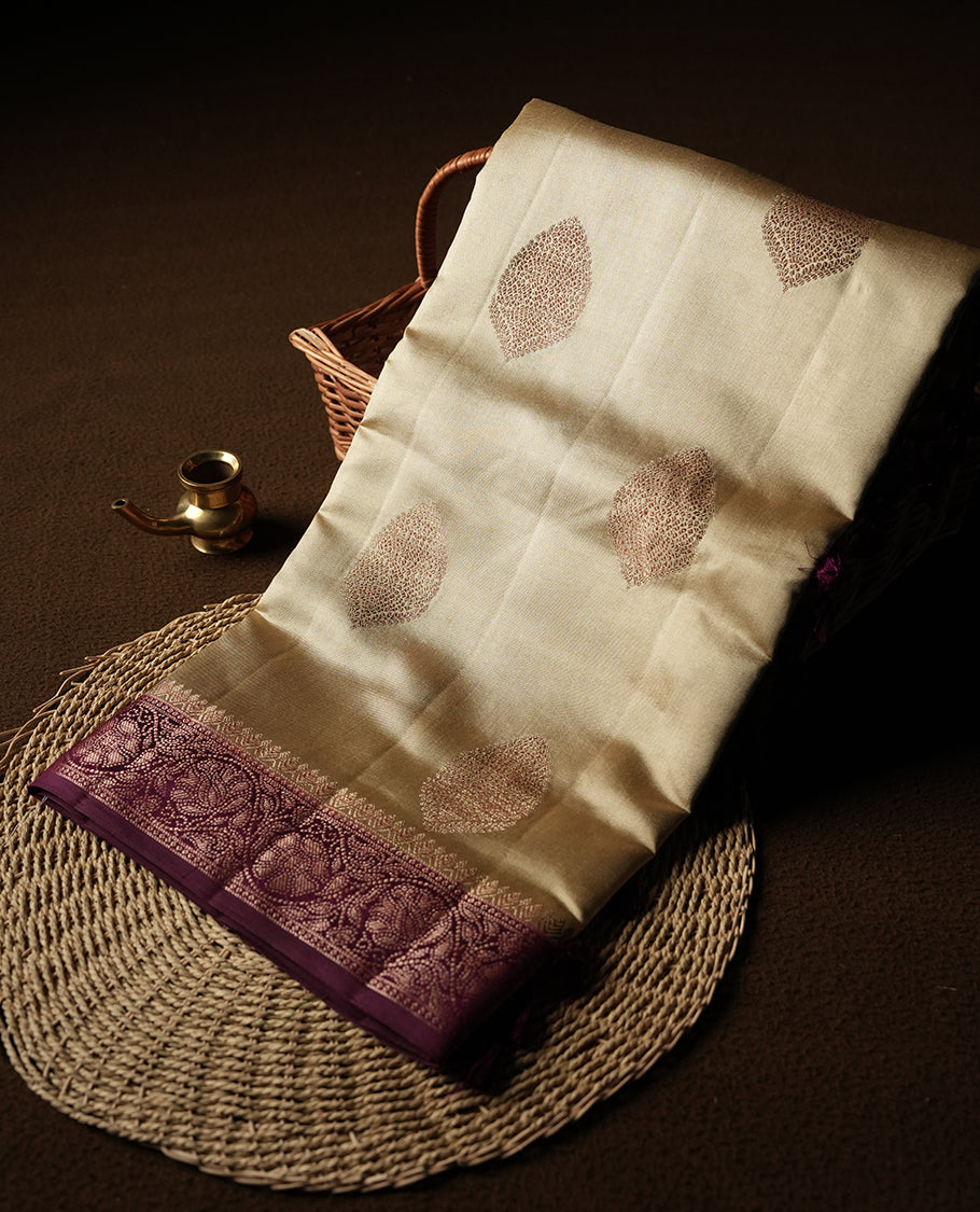 Cream Colour Banaras Soft Silk Saree with Gold Zari Oval Leaf Buttas, Intricate Multi-Layered Floral Border, Grand Pallu with Intricate Brocade Motifs in Deep Maroon and Gold Tones, and Matching Blouse.