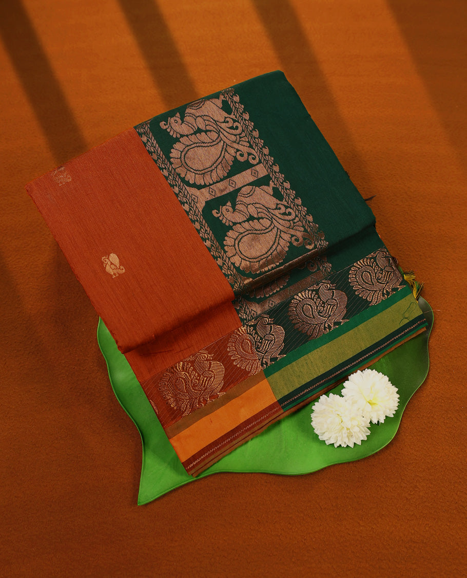 Rust Orange colour mixed Kalyani cotton saree featuring Copper zari Peacock motifs throughout the body, paired with Bottle Green colour traditional border and pallu, with matching blouse