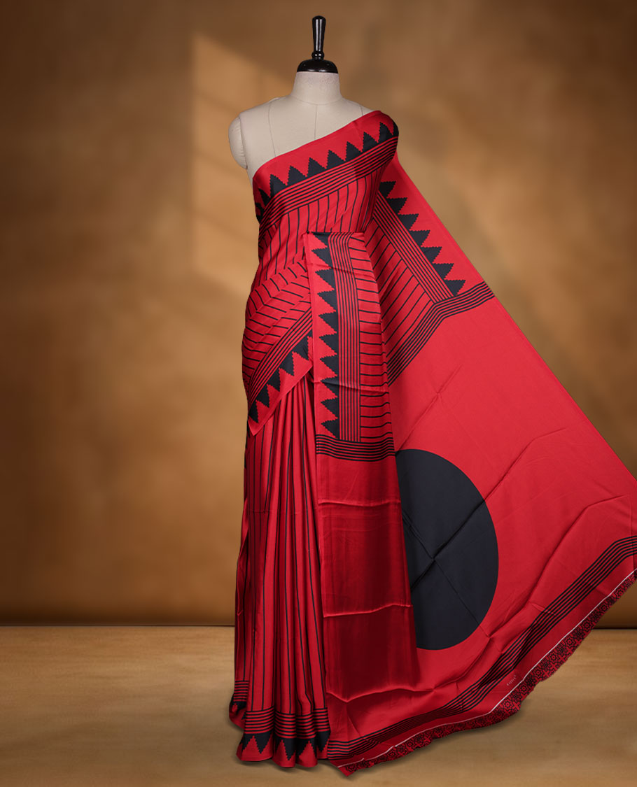 Red colour semi modal saree featuring black colour geometric borders and fine stripe detailing, finished with an oversized circular motif on the pallu, paired with a matching printed blouse.