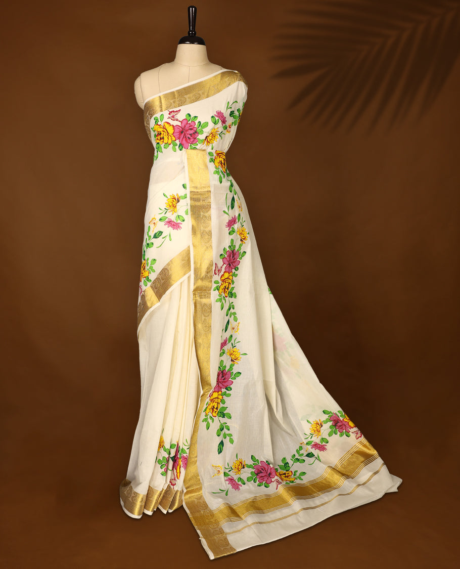 Elegant off white cotton Kerala saree adorned with hand painted floral motifs in pink, yellow, and green, beautifully accented by a rich golden zari border and pallu paired with matching blouse