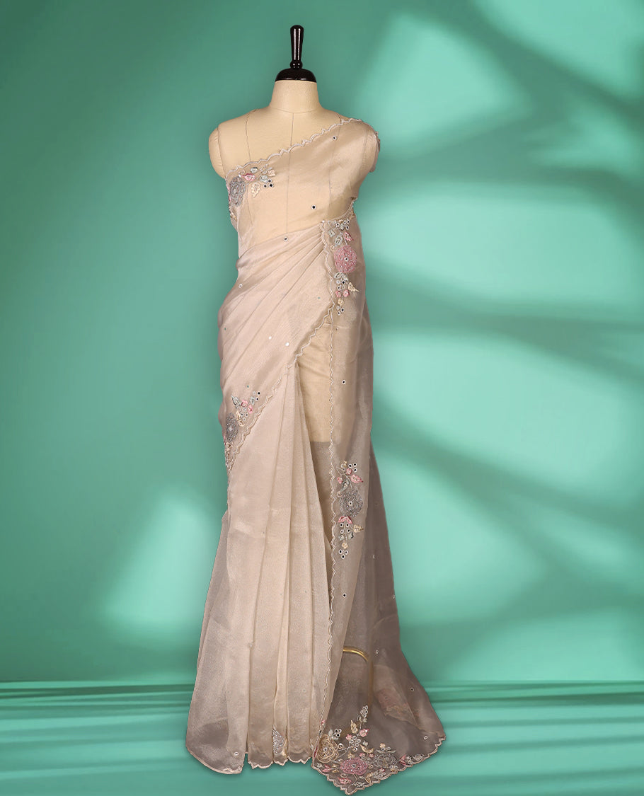 Beige Colour organza saree featuring delicate floral embroidery with applique, bead, mirror, and thread detailing on the body and scalloped border, lending a soft feminine charm, paired with a matching blouse.