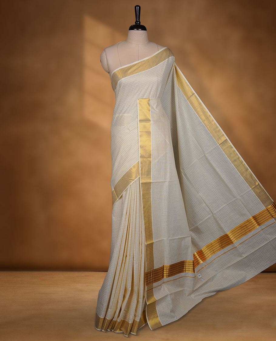 Off-white colour cotton Kerala saree featuring a subtle gold checkered weave, highlighted with a rich traditional gold kasavu border along the pallu and pleats, and paired with a matching blouse.