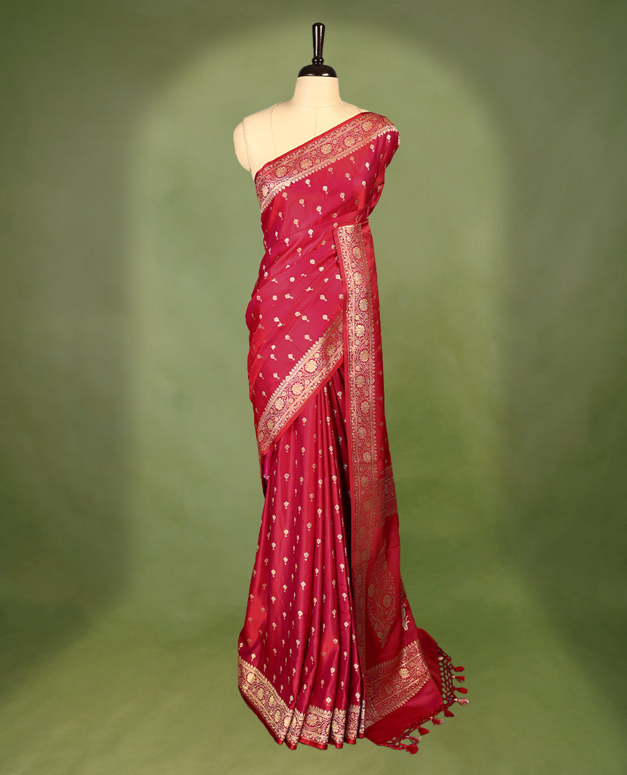 Pinkish red colour semi Banaras satin crepe saree featuring gold zari woven flower motifs across the body, complemented by an intricately designed traditional banarasi border and richly woven pallu, paired with a matching blouse piece.