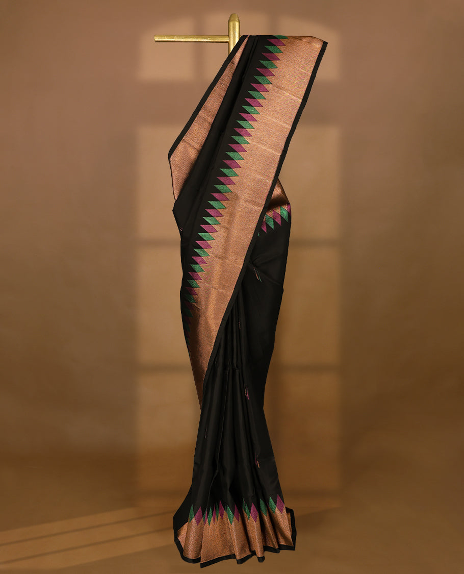 Solid black Colour traditional Kancheepuram silk saree adorned with antique zari motifs on the body, featuring a border with zigzag triangular patterns in alternating green and magenta over a coppery gold zari weave, and paired with a matching blouse.