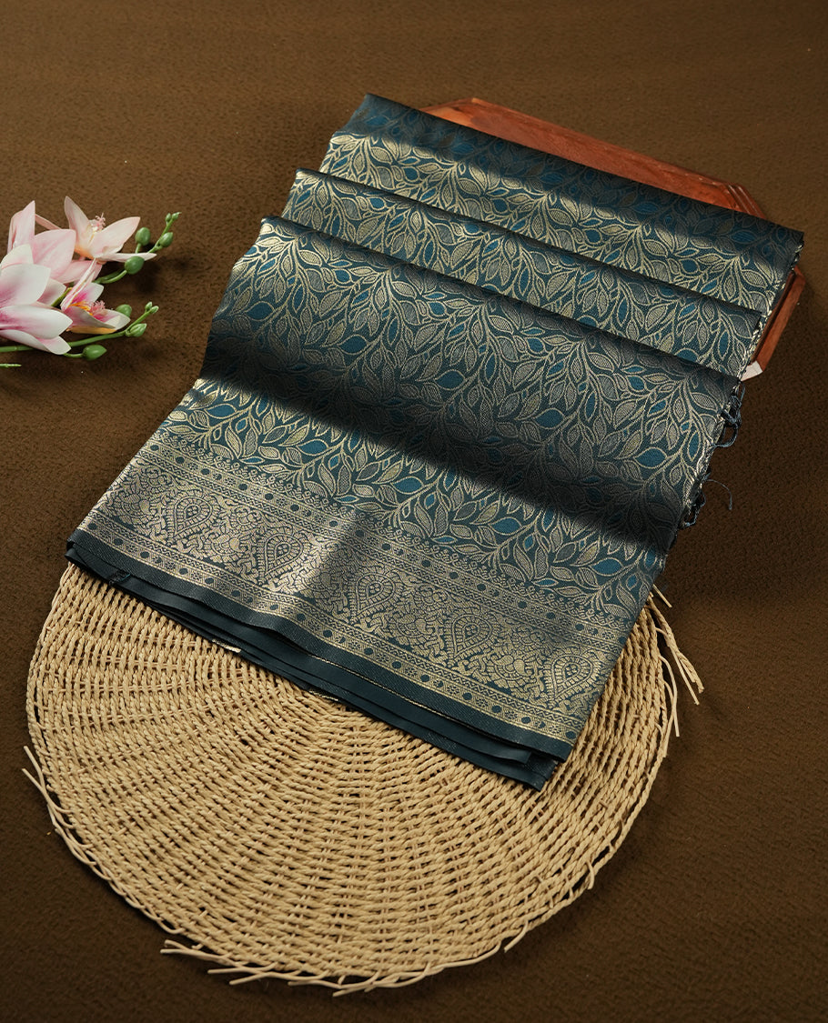 Dark Teal Green colour semi soft silk saree featuring an all-over woven leaf pattern in gold colour, finished with a matching border adorned with detailed paisley and floral motifs, and a matching blouse piece.