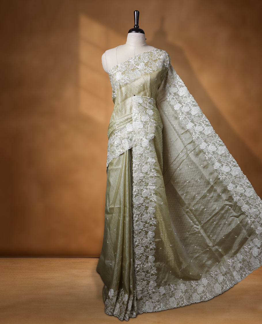 Pastel olive colour organza designer saree featuring a subtle self patterned body and an intricately scalloped floral thread embroidered border all around, complemented by a coordinating embroidered pallu and paired with a matching blouse.
