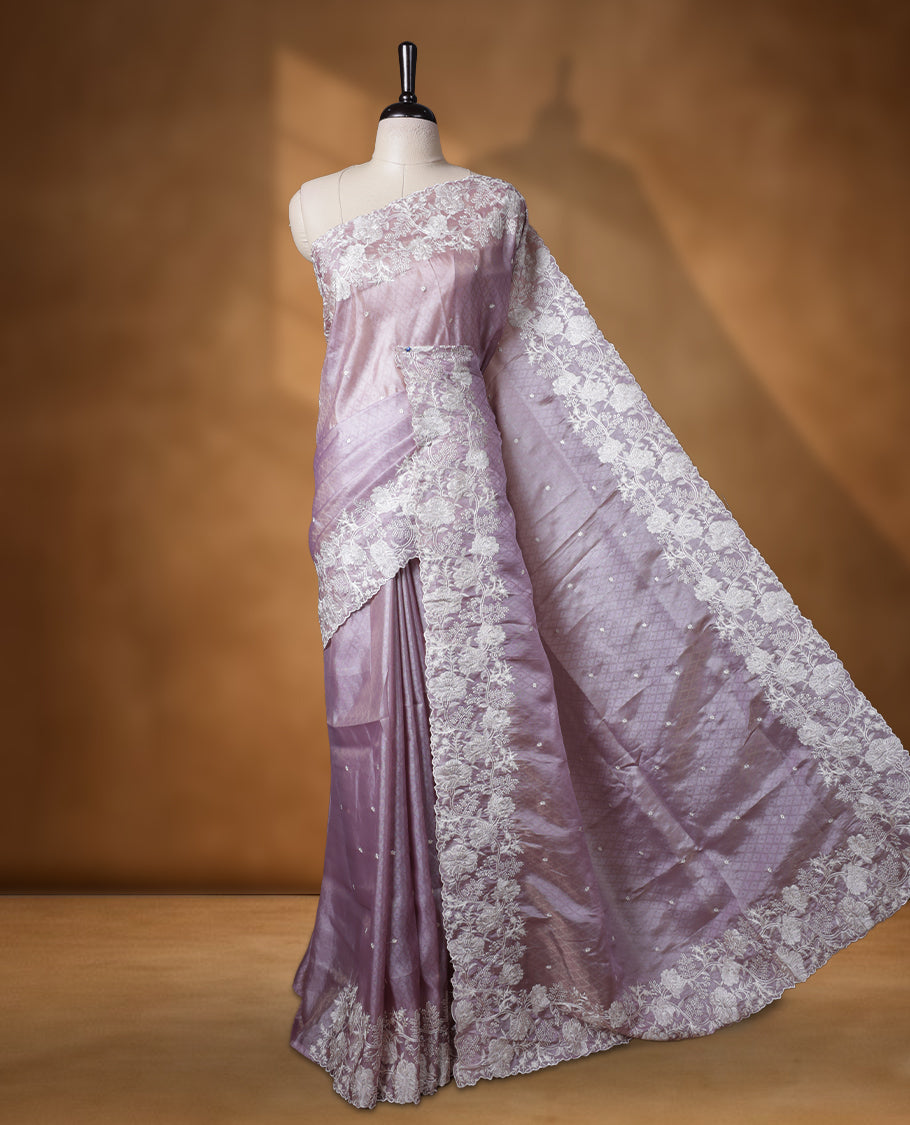 Lavender colour organza designer saree featuring a subtle self patterned body and an intricately scalloped floral thread embroidered border all around, complemented by a coordinating embroidered pallu and paired with a matching blouse.