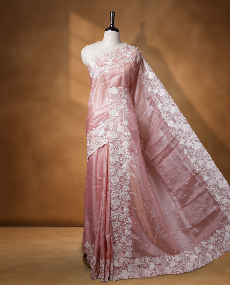 Pastel peach colour organza designer saree featuring a subtle self patterned body and an intricately scalloped floral thread embroidered border all around, complemented by a coordinating embroidered pallu and paired with a matching blouse.