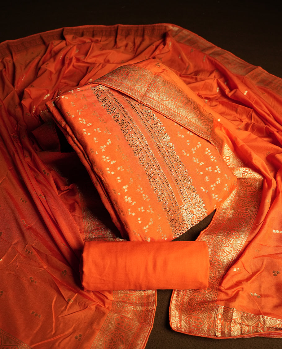 Orange colour churidar material adorned with small golden stone work on the neckline, featuring intricate zari floral motifs on the body, rich zari woven border and dupatta, paired with matching plain bottom.
