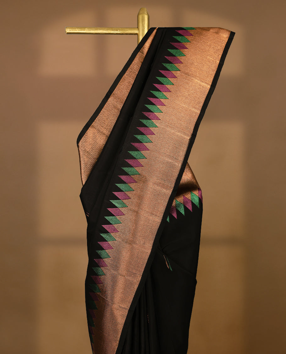 Solid black Colour traditional Kancheepuram silk saree adorned with antique zari motifs on the body, featuring a border with zigzag triangular patterns in alternating green and magenta over a coppery gold zari weave, and paired with a matching blouse.