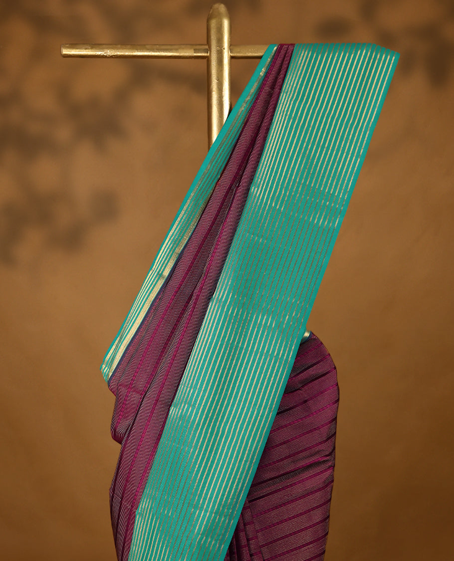 Grape coloured soft silk saree, adorned with golden zari stripes across the body, paired with a contrasting aqua-coloured border embellished with intricate golden zari work, paired with a matching contrast blouse