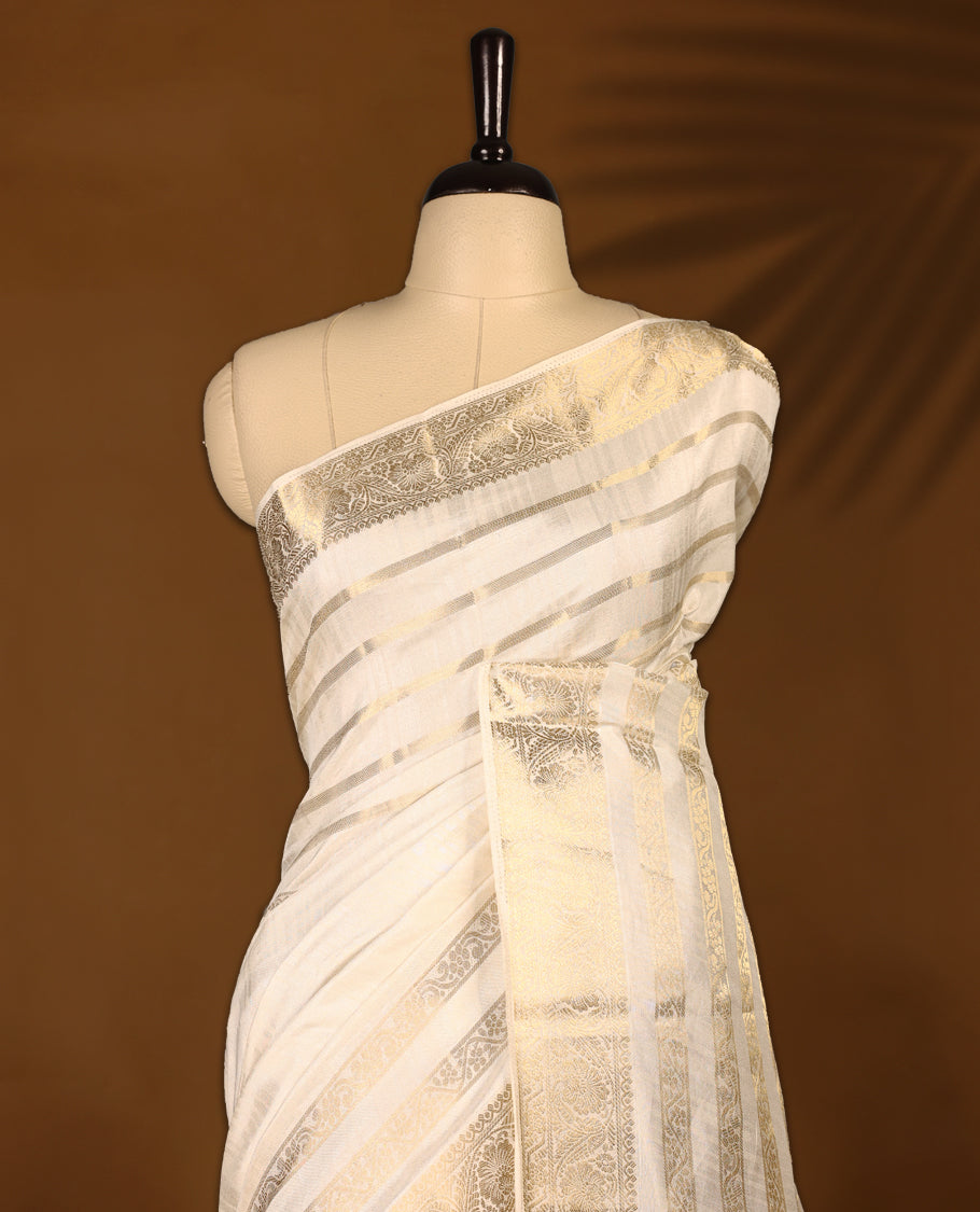 Ivory white Colour Tussar Kerala saree with a sheer, smooth body and soft golden sheen, Accented with fine zari stripes and an intricate floral zari border,Comes paired with a coordinated matching blouse piece.