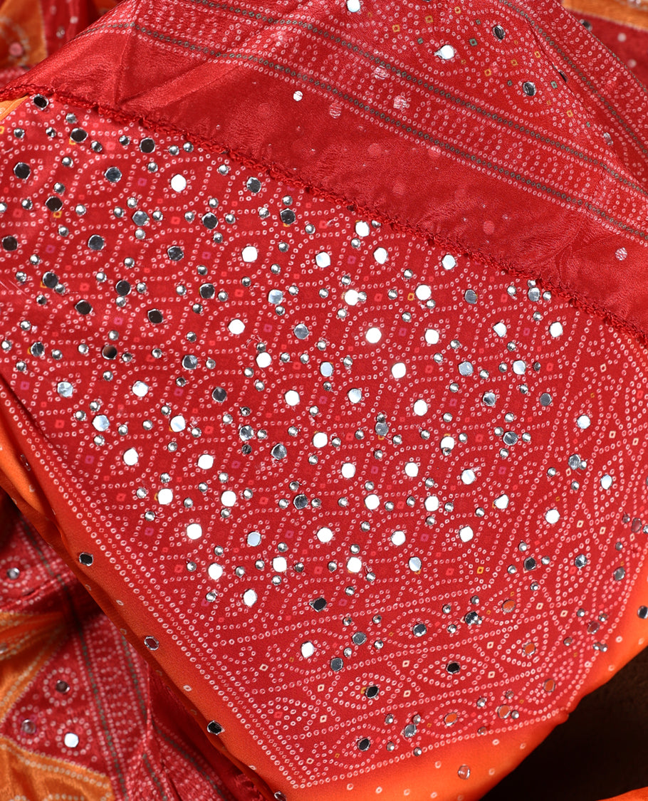 Red and orange colour churidhar material with intricate bandhani patterns and dazzling mirror work, paired with a plain matching bottom and vibrant dupatta, perfect for festive occasions and cultural celebrations.