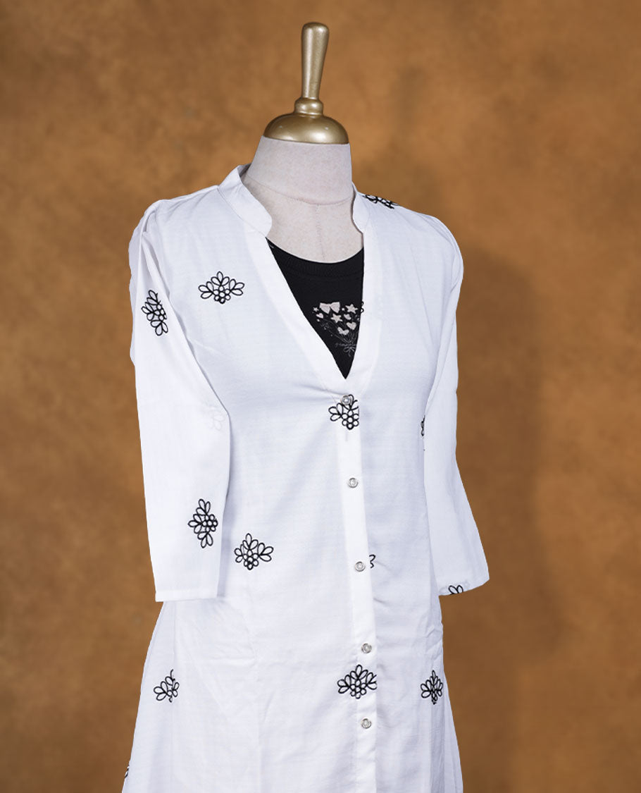 White embroidered shirt dress with band collar, contrast black inner layer at neckline, front button placket, three-quarter sleeves, and intricate black border embroidery at hem