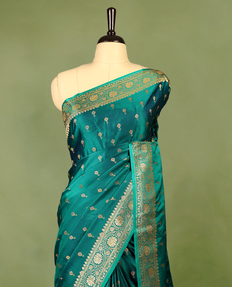Deep sea green colour semi Banaras satin crepe saree featuring gold zari woven flower motifs across the body, complemented by an intricately designed traditional banarasi border and richly woven pallu, paired with a matching blouse piece.