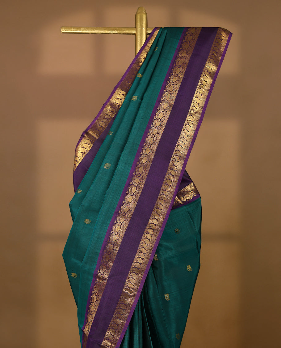 Deep teal blue Colour pure Kancheepuram silk saree with small peacock and elephant motifs, featuring a deep purple border with arched floral and paisley gold zari patterns, paired with a matching blouse.
