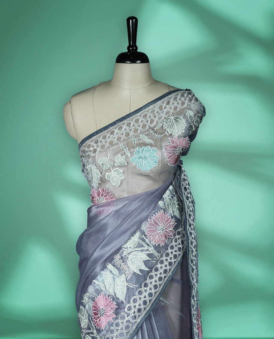 Grey colour organza designer saree featuring delicate pastel floral embroidery in pink, mint green, and white across the body, with a scalloped border accentuated by floral threadwork, and a gracefully embroidered pallu paired with matching blouse.