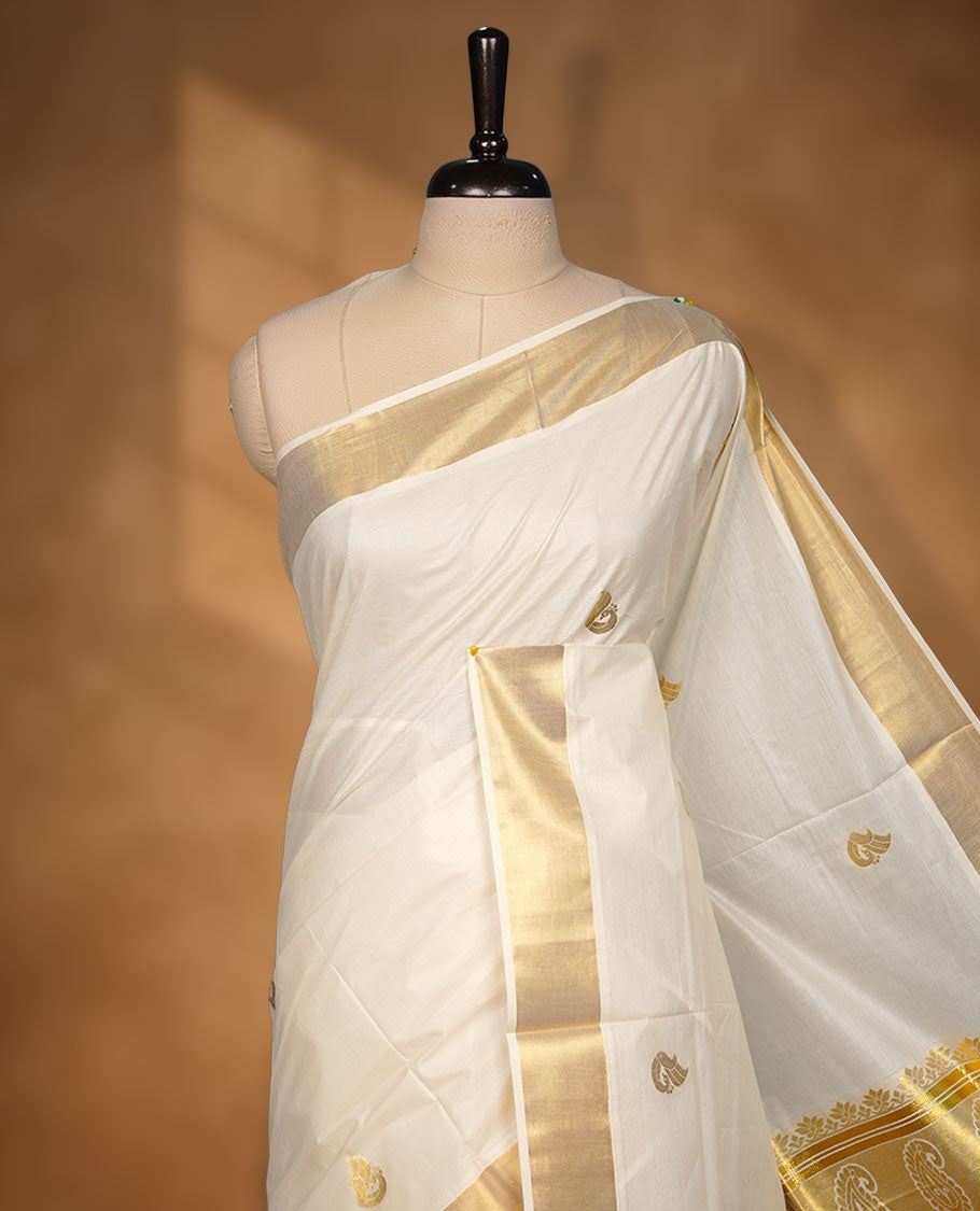Off-white cotton Kerala saree with subtle gold zari buttas, a broad kasavu border, and a richly woven traditional pallu with paisley motifs. Paired with a matching plain blouse