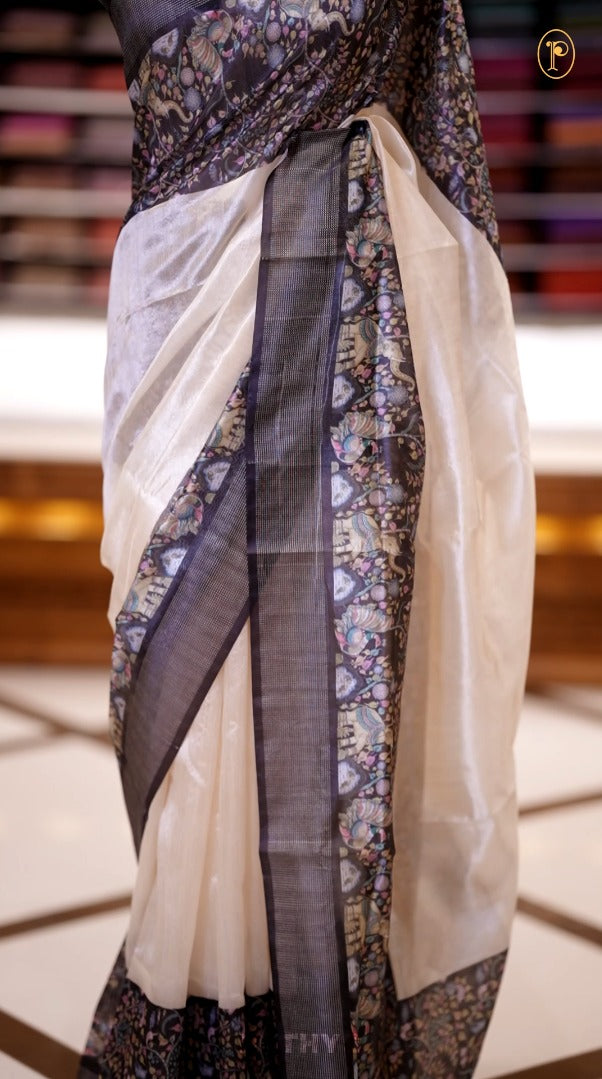 Beige colour semi tussar silk saree featuring with a soft kalamkari & floral prints, accented with a rich black and silver border and elegant printed pallu, paired with a black colour printed with border blouse.