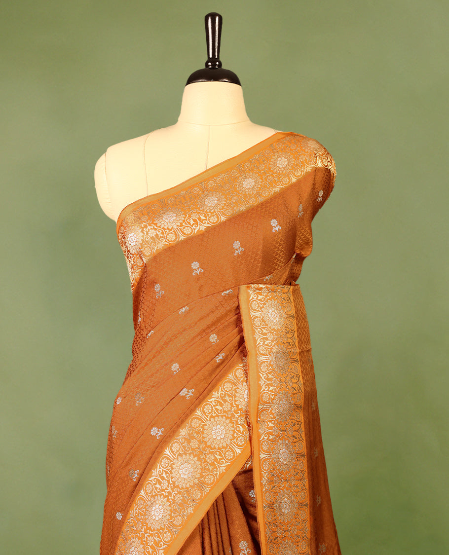Burnt orange colour pure Banaras crepe saree showcases elegant silver zari Flower Motifs all over the body, a traditional Banarasi gold zari border, a rich intricately woven pallu, and comes with a matching blouse.