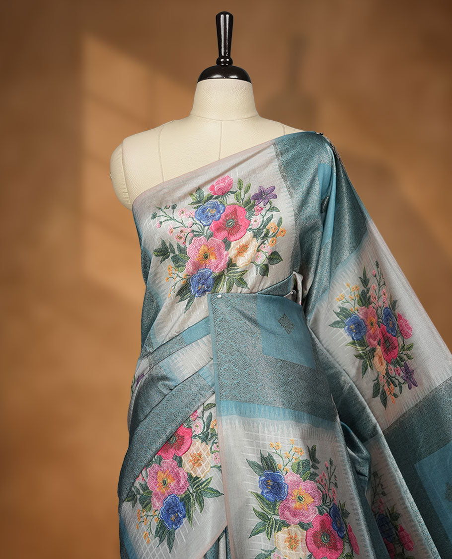 Blue color semi tussar Banarasi saree featuring vibrant multi-color floral prints across the body, with geometric zari patterns, and a matching blouse detailed with silver woven buttas.