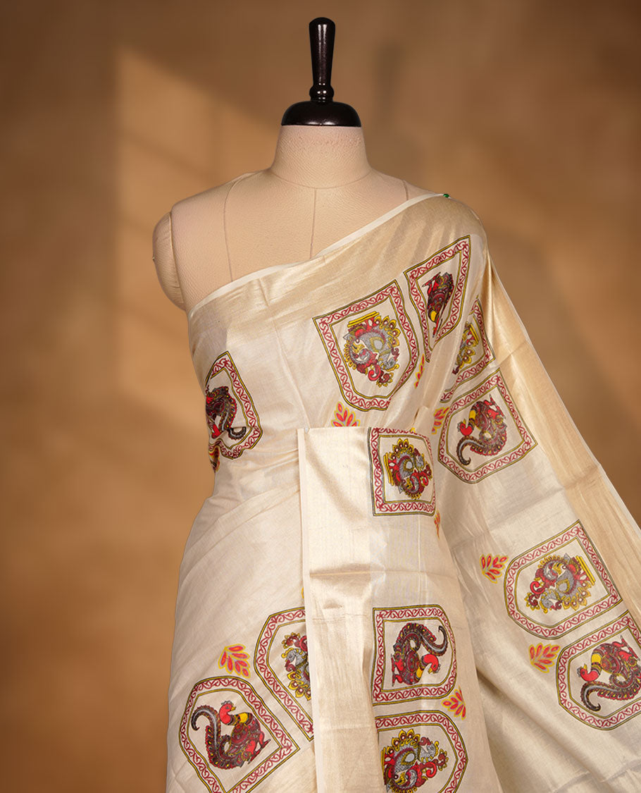 Off-white colour tissue Kerala saree, featuring a rich gold kasavu border and traditional Kalamkari-inspired peacock and elephant motifs across the body and pallu. Lightweight, breathable, and paired with a matching blouse.