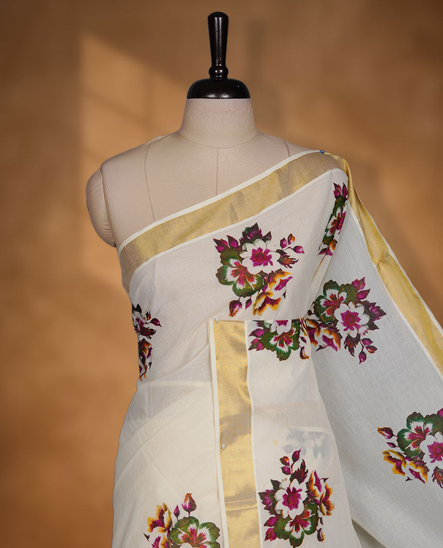 Off-white colour cotton Kerala saree with a classic gold kasavu border, adorned with multi colour floral motifs across the body and pallu, and paired with a matching blouse.