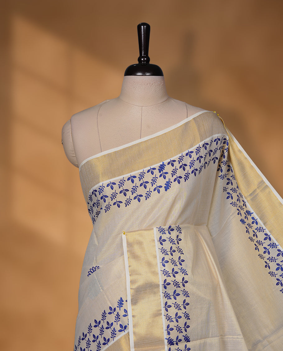 Off-white colour tissue Kerala saree with a rich gold kasavu border, adorned with delicate royal blue floral motifs across the body and pallu, paired with a matching blouse.