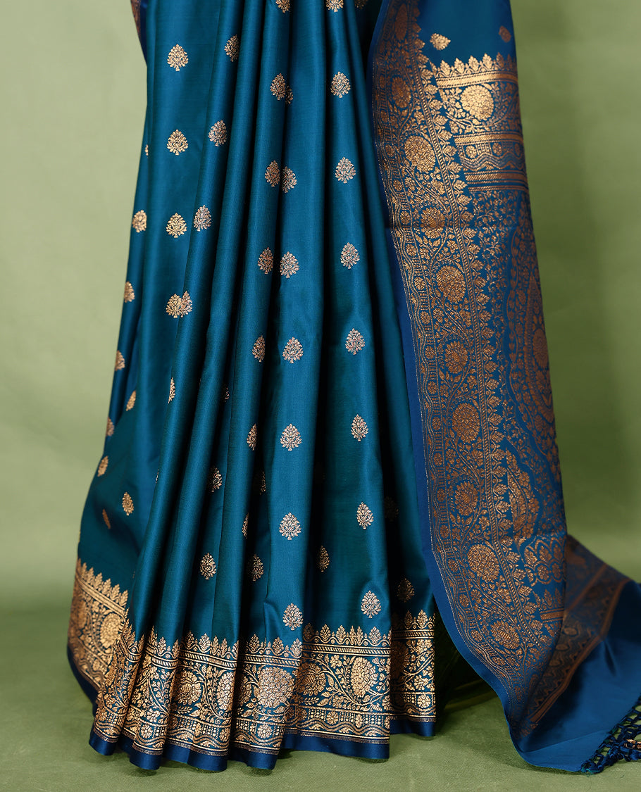Peacock blue colour semi Banaras satin crepe saree featuring gold zari woven floral motifs across the body, complemented by an intricately designed traditional banarasi border and richly woven pallu, paired with a matching blouse piece.