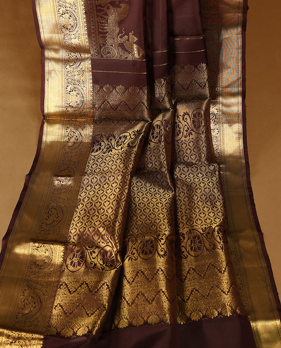 Dark brown colour pure traditional Kancheepuram silk saree adorned with golden Floral motifs on the body, featuring an exquisite antique zari border, paired with a contrasting blouse.
