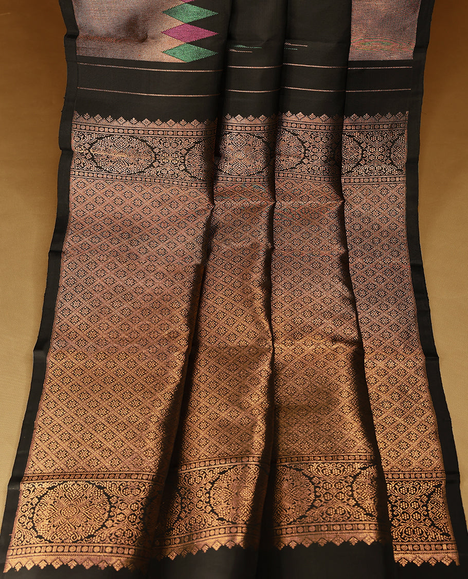 Solid black Colour traditional Kancheepuram silk saree adorned with antique zari motifs on the body, featuring a border with zigzag triangular patterns in alternating green and magenta over a coppery gold zari weave, and paired with a matching blouse.
