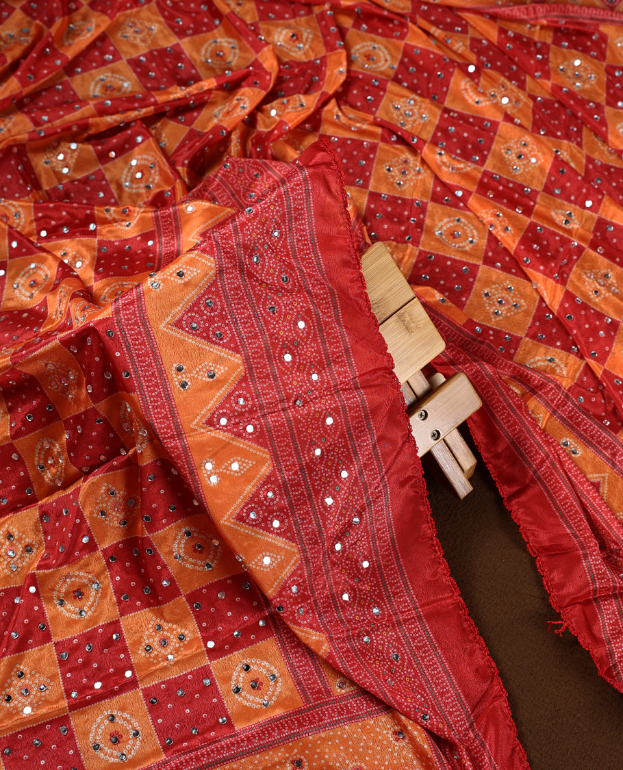 Red and orange colour churidhar material with intricate bandhani patterns and dazzling mirror work, paired with a plain matching bottom and vibrant dupatta, perfect for festive occasions and cultural celebrations.