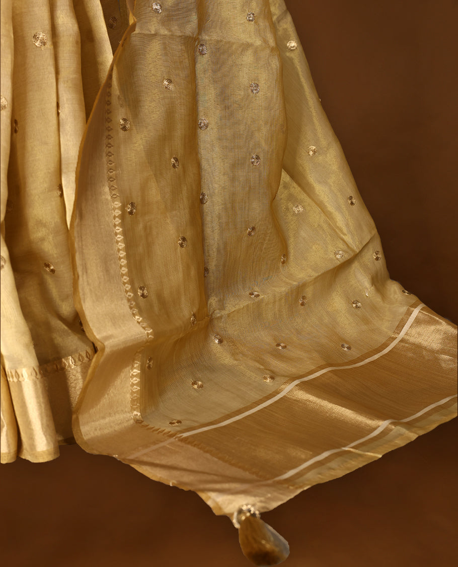 Light gold colour Organza Kerala saree featuring a luminous body with a subtle golden sheen, enhanced by a sleek zari border and scattered sequin like floral motifs, Paired elegantly with a matching blouse piece.