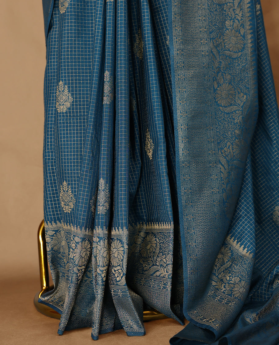 Blue coloured semi Banaras georgette saree featuring golden Zari checks with floral motifs on the body, complemented by a self coloured traditional border with floral kodi design and a matching blouse.