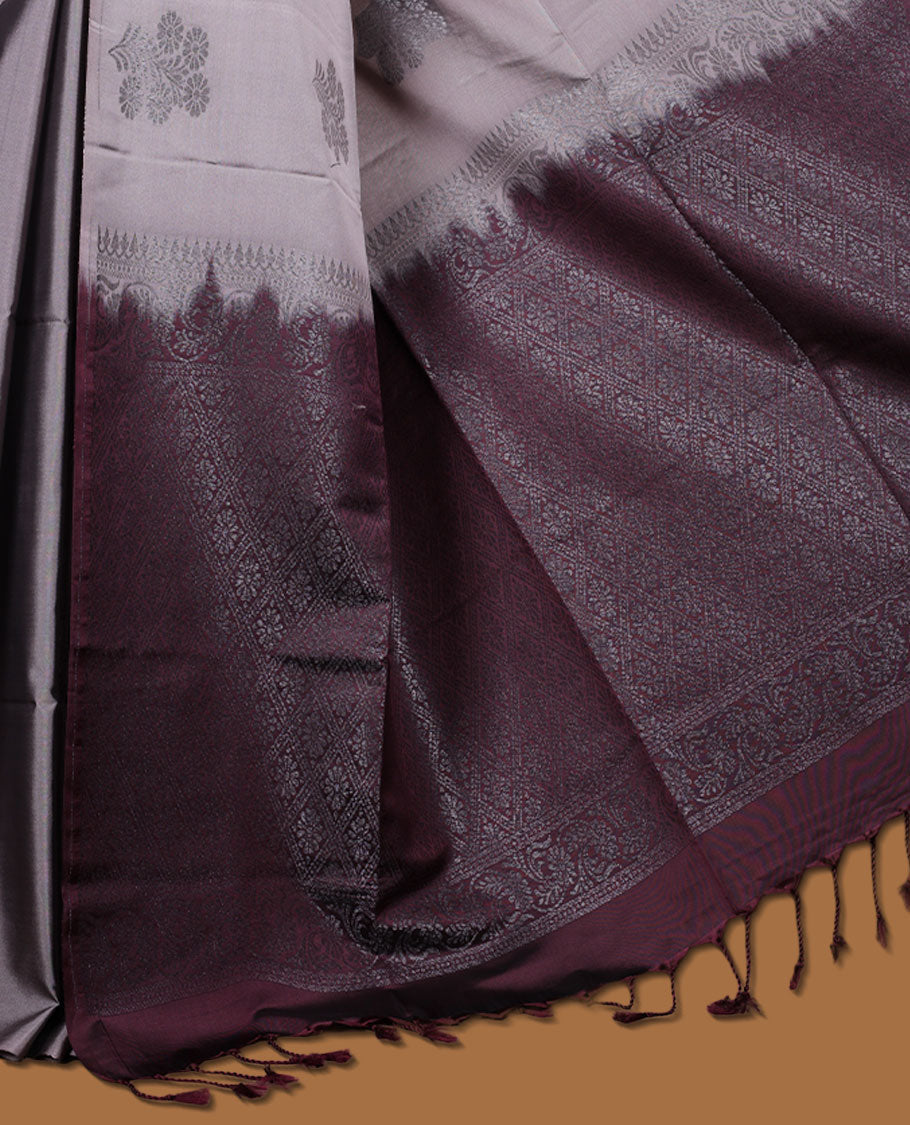 Reddish gray colour semi soft silk saree adorned with delicate woven floral motifs on the body, complemented by a contrast deep maroon colour woven pallu with intricate detailing and tassel accents without border & Comes with a matching blouse piece.