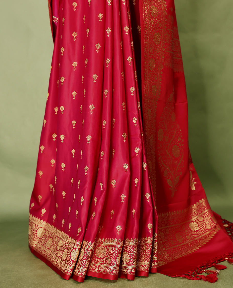 Pinkish red colour semi Banaras satin crepe saree featuring gold zari woven flower motifs across the body, complemented by an intricately designed traditional banarasi border and richly woven pallu, paired with a matching blouse piece.