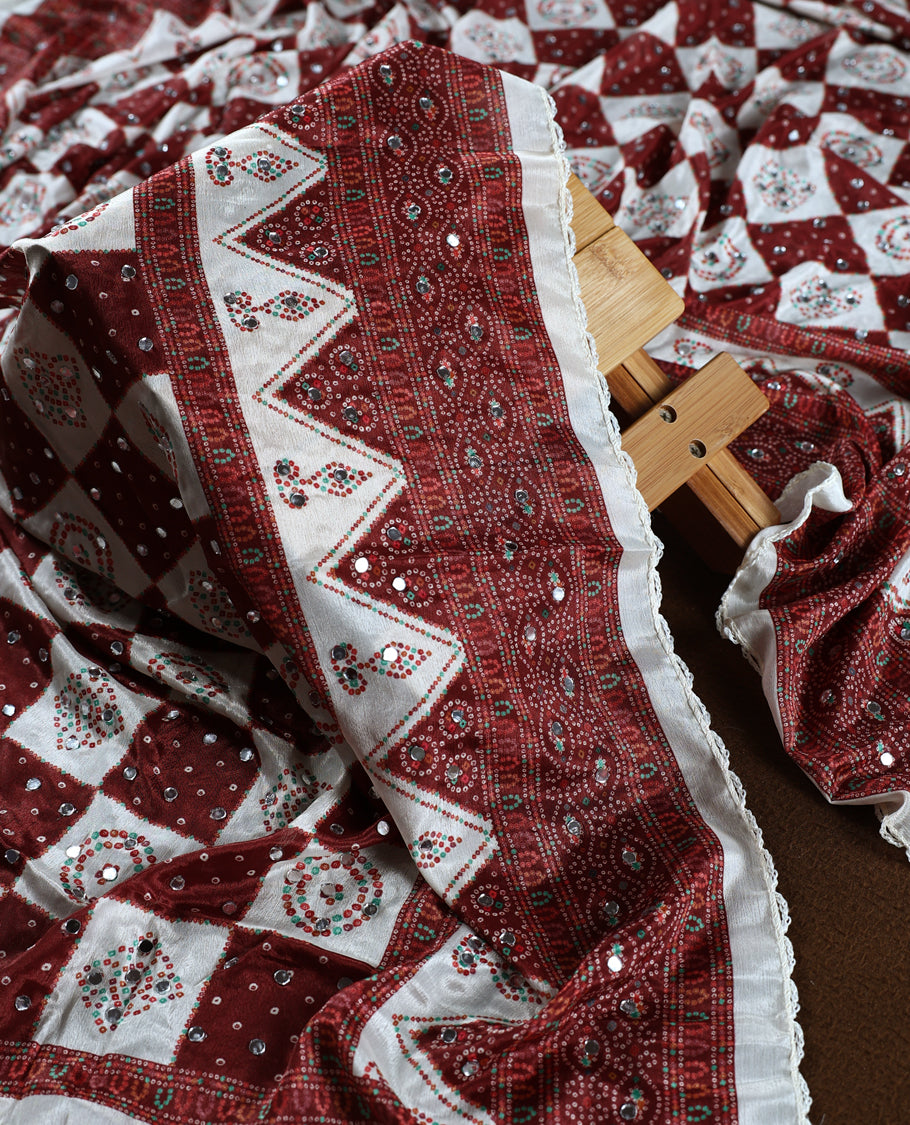 Off white colour churidhar material adorned with mirror work and maroon colour traditional bandhani patterns, paired with a green and maroon colour dotted off white top, plain bottom, and an elegant matching dupatta.
