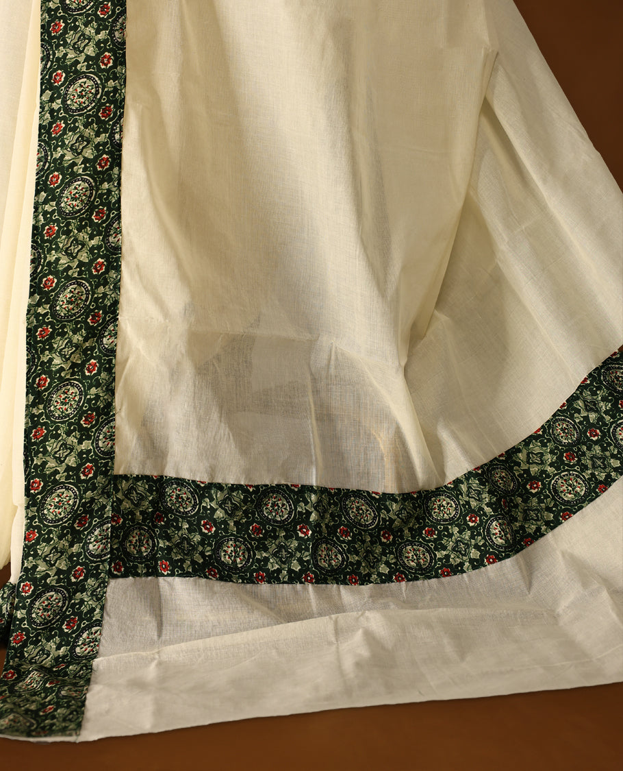 off white colour cotton Kerala saree adorned with a deep green border featuring intricate floral motifs in red and cream, paired with a matching printed blouse for a refined traditional look