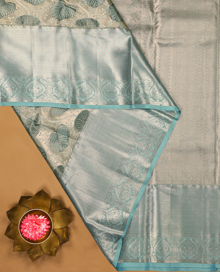 Silver Colour Kancheepuram tissue silk saree featuring intricate silver and blue zari leaf motifs on the body, gracefully complemented by a light blue traditional peacock and floral design border, and paired with a matching blouse.