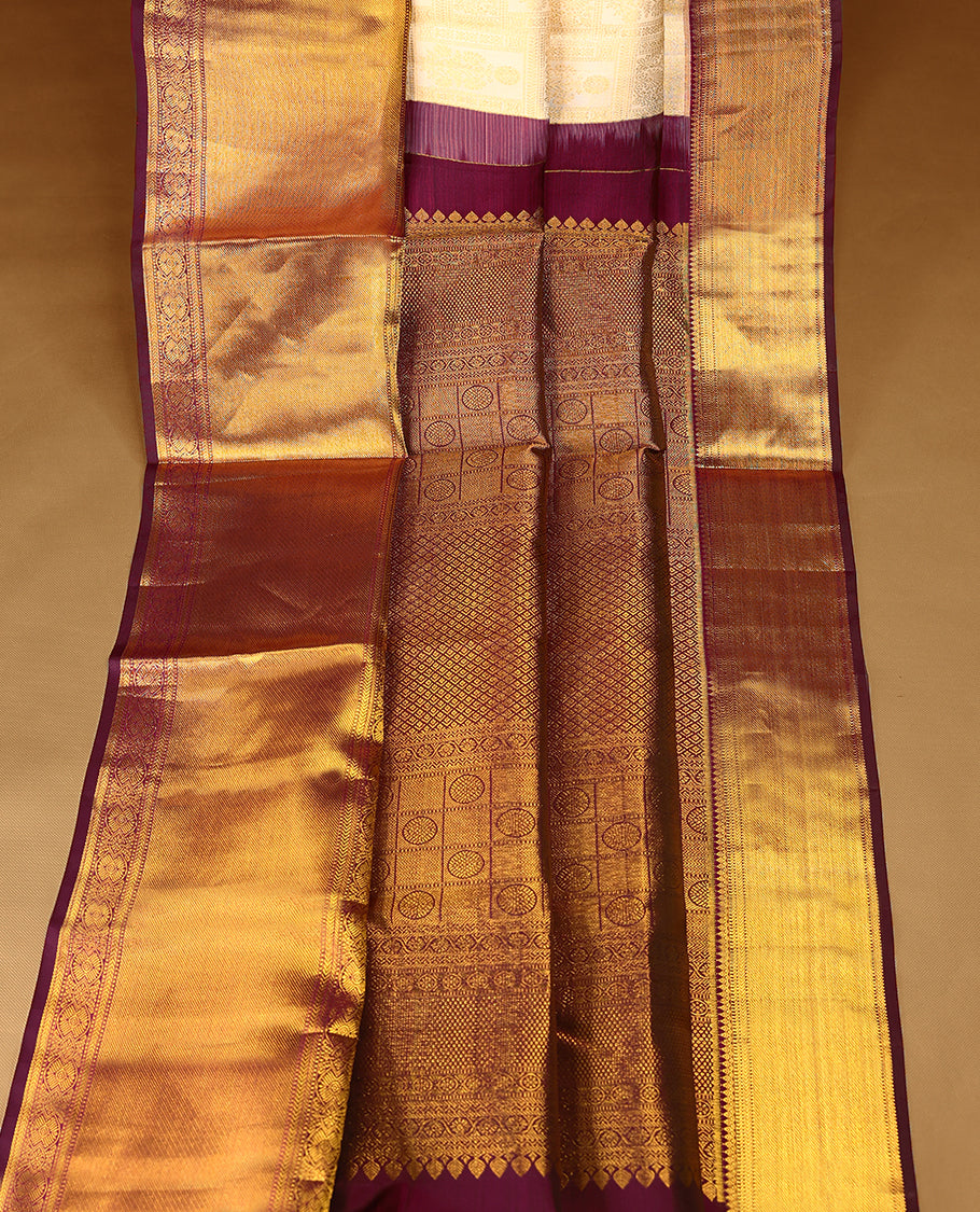 Off-white Colour traditional Kancheepuram silk saree adorned with antique zari floral motifs arranged in a symmetric grid on the body, featuring a contrasting border with floral, peacock, and chakram motifs, and paired with a matching blouse.