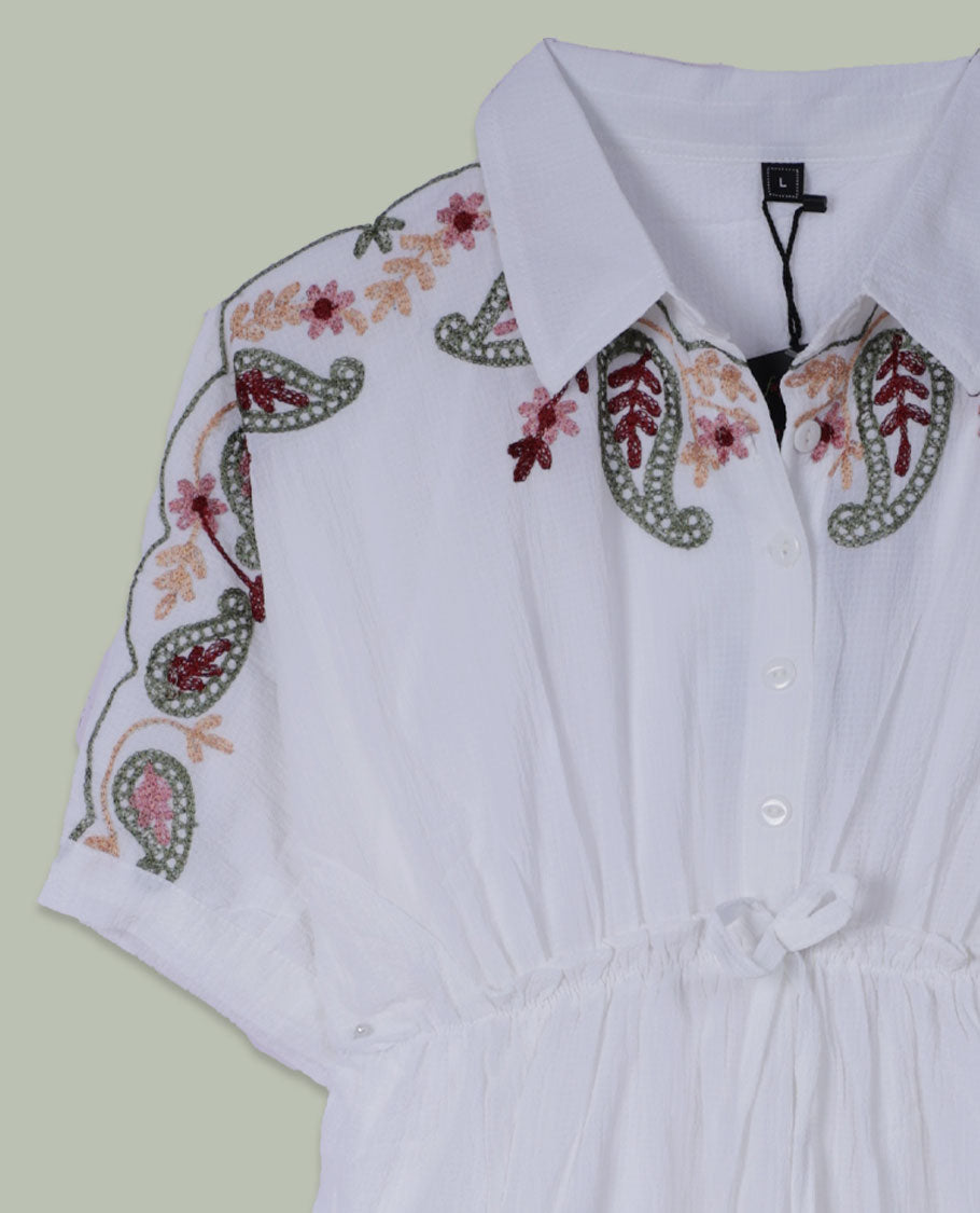 White short tunic top with collared neckline, short sleeves, multicolour paisley and floral embroidery on shoulders and hem, and a drawstring waist with long tasseled ties.