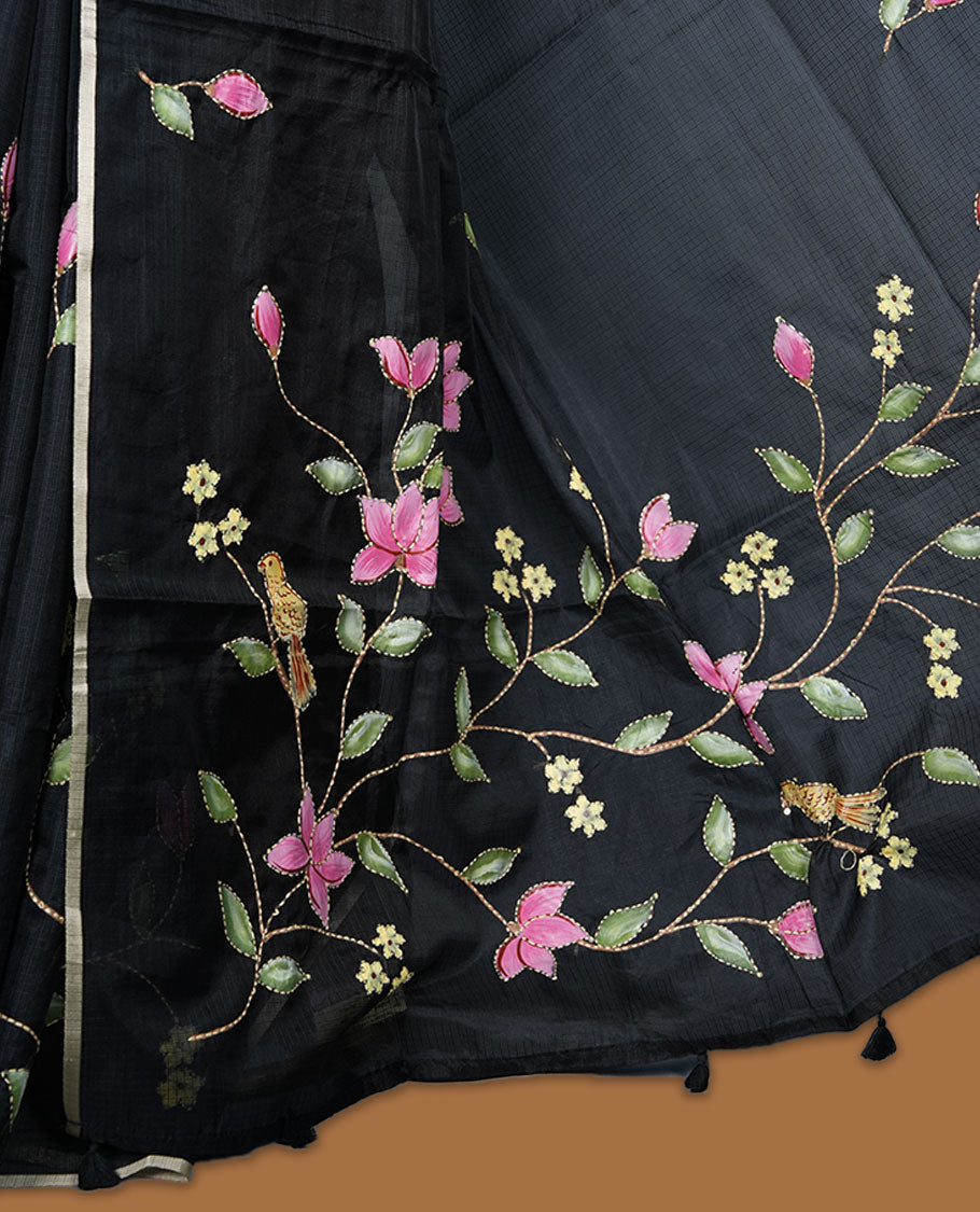Black colour semi tusser silk saree adorned with fine floral embroidery spread across the body, highlighted by subtle contrast accents along the border, paired with a matching blouse.