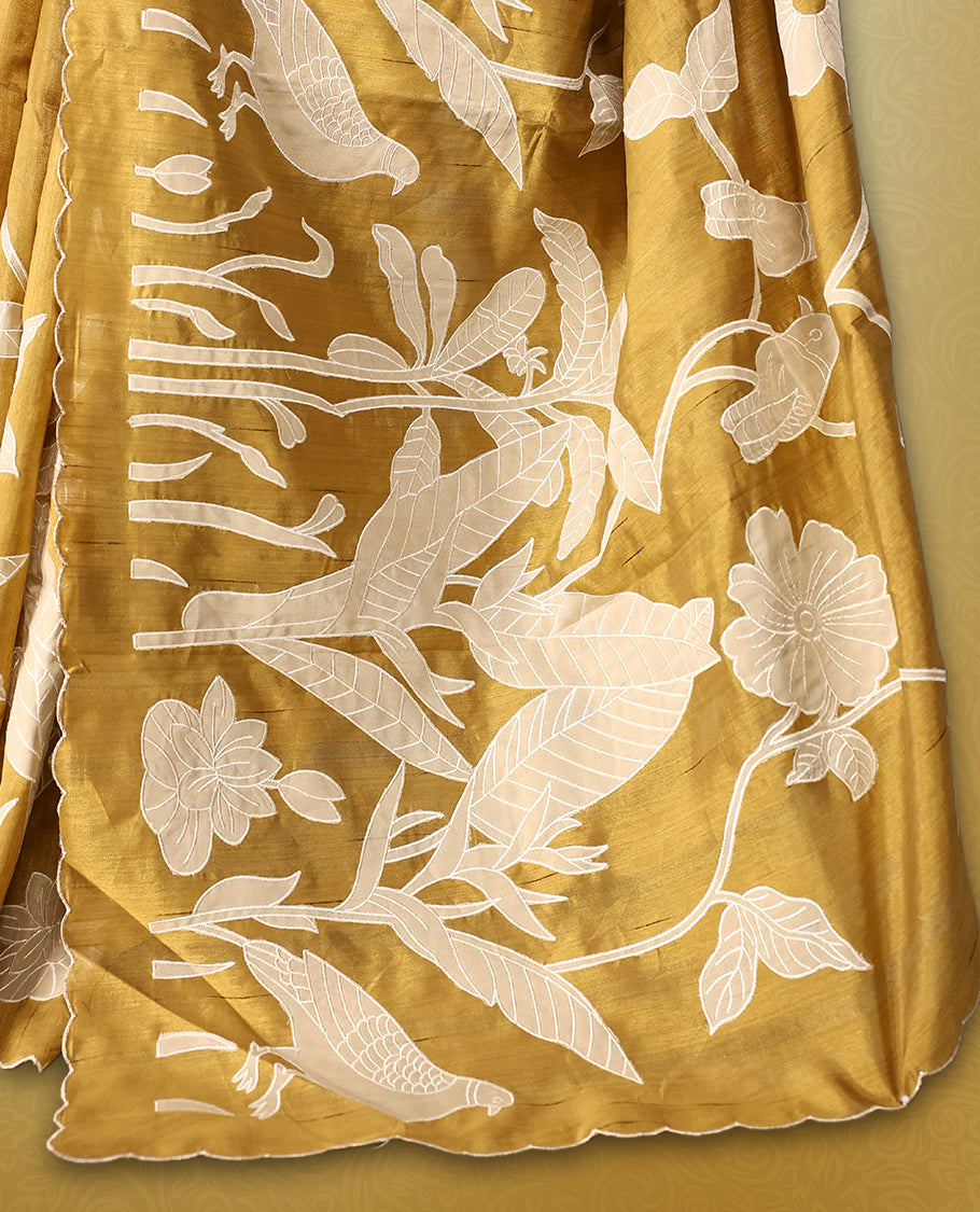 Golden Brown colour semi tussar saree adorned with elegant white applique work in leaf and floral designs across the body and border, paired with a matching blouse.
