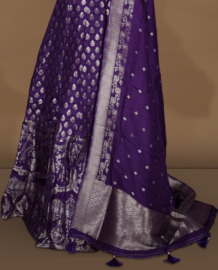 Purple colour Banarasi lehenga set with a stitched flared lehenga featuring all‑over zari butti motifs and rich woven border, paired with an unstitched floral zari blouse piece and matching dupatta with zari border and tasselled edges.
