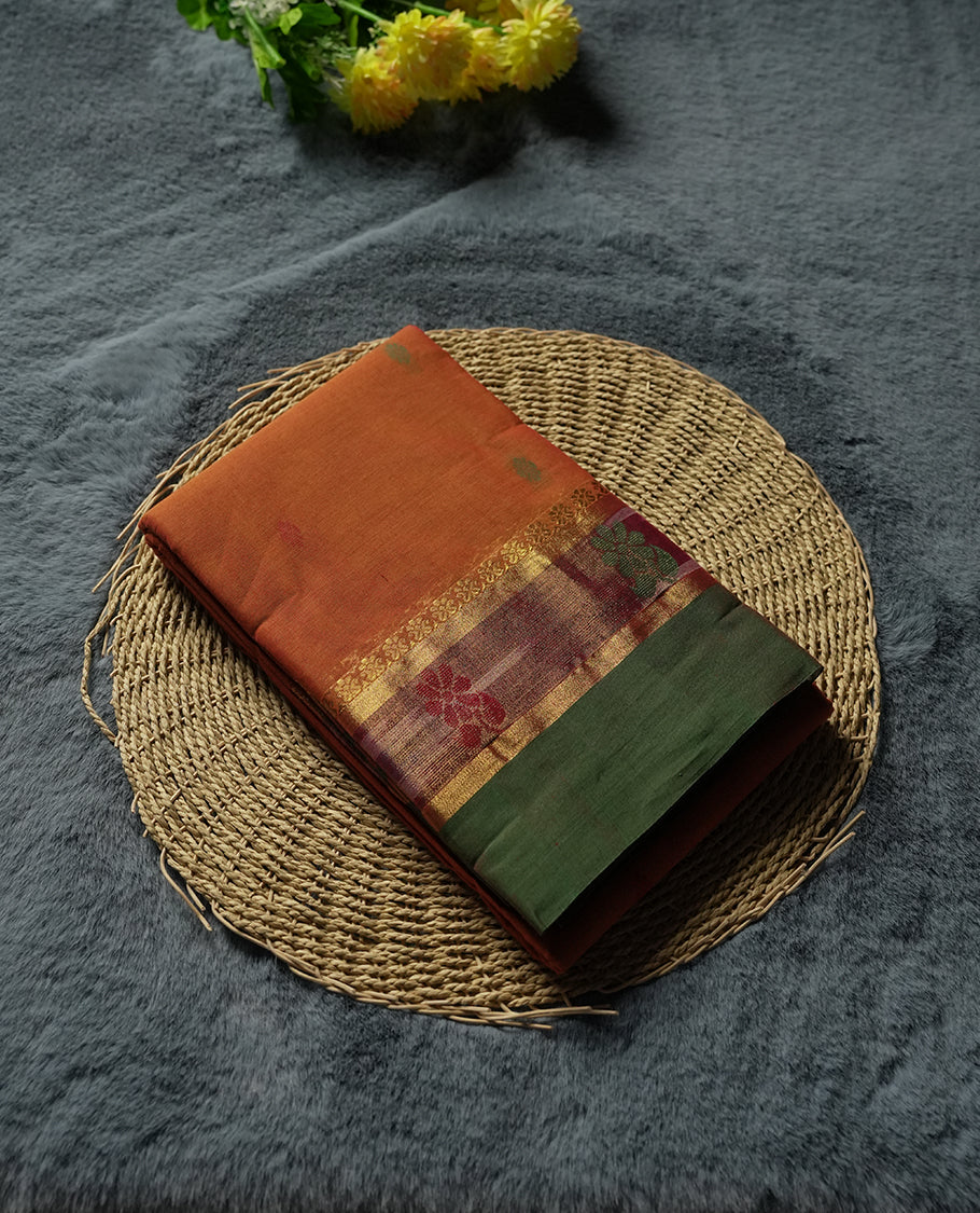 Burnt Orange colour Silk Cotton Saree with Contrast Green Zari Border featuring Ikat-inspired floral panels, Small Dual-Tone Buttas on the Body, Paisley-Woven Pallu, and a Coordinated Matching Blouse Piece