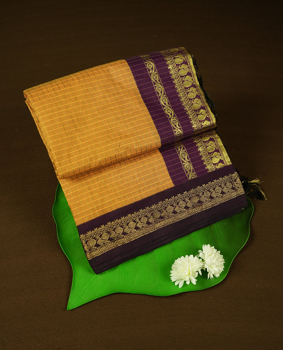 Mustard Yellow coloured mixed Kalyani cotton saree featuring golden zari checks on the body, complemented by a dark purple coloured border and pallu adorned with golden zari patterns, Paired with a matching blouse.