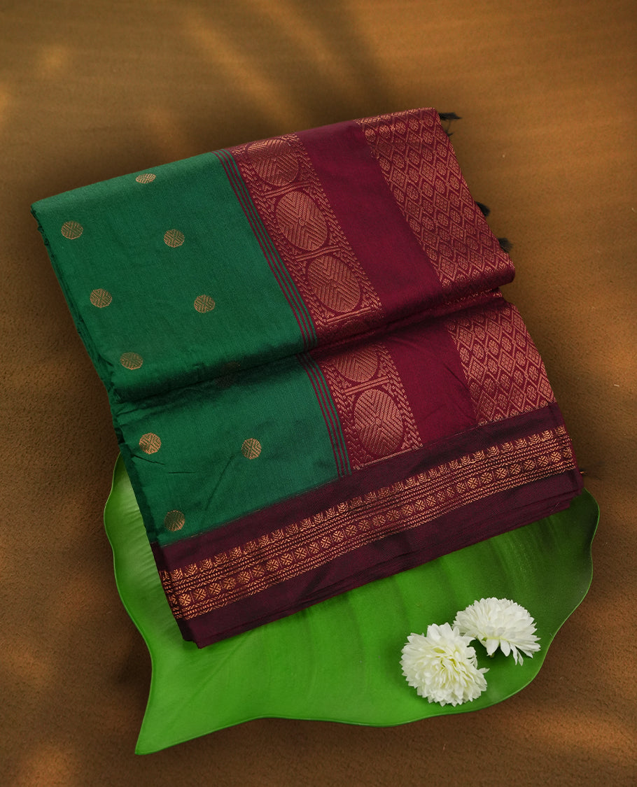 Green colour Kalyani Cotton saree with golden motifs, paired with a contrasting maroon border featuring intricate zari work & Comes with a matching blouse piece.