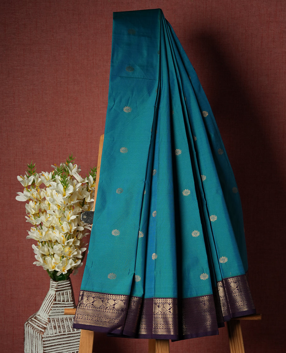 Teal Blue Colour Semi Silk Saree with Golden Peacock Butta Motifs, Contrasting Purple Golden Zari Pallu & Border, Traditional Leaf Designs, and Matching Blouse Piece