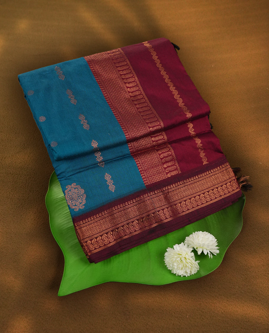 Blue colour Kalyani Cotton saree in deep maroon with golden floral motifs, paired with a contrasting maroon border featuring intricate zari work & Comes with a matching blouse piece.