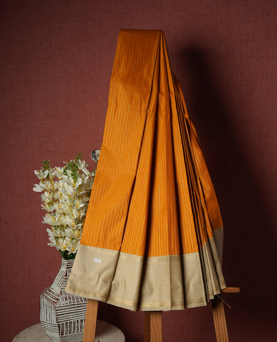 Burnt orange colour Semi Silk Saree with gold zari striped patterns on the body, paired with a contrasting cream colour traditional pallu and border, and a matching blouse piece.